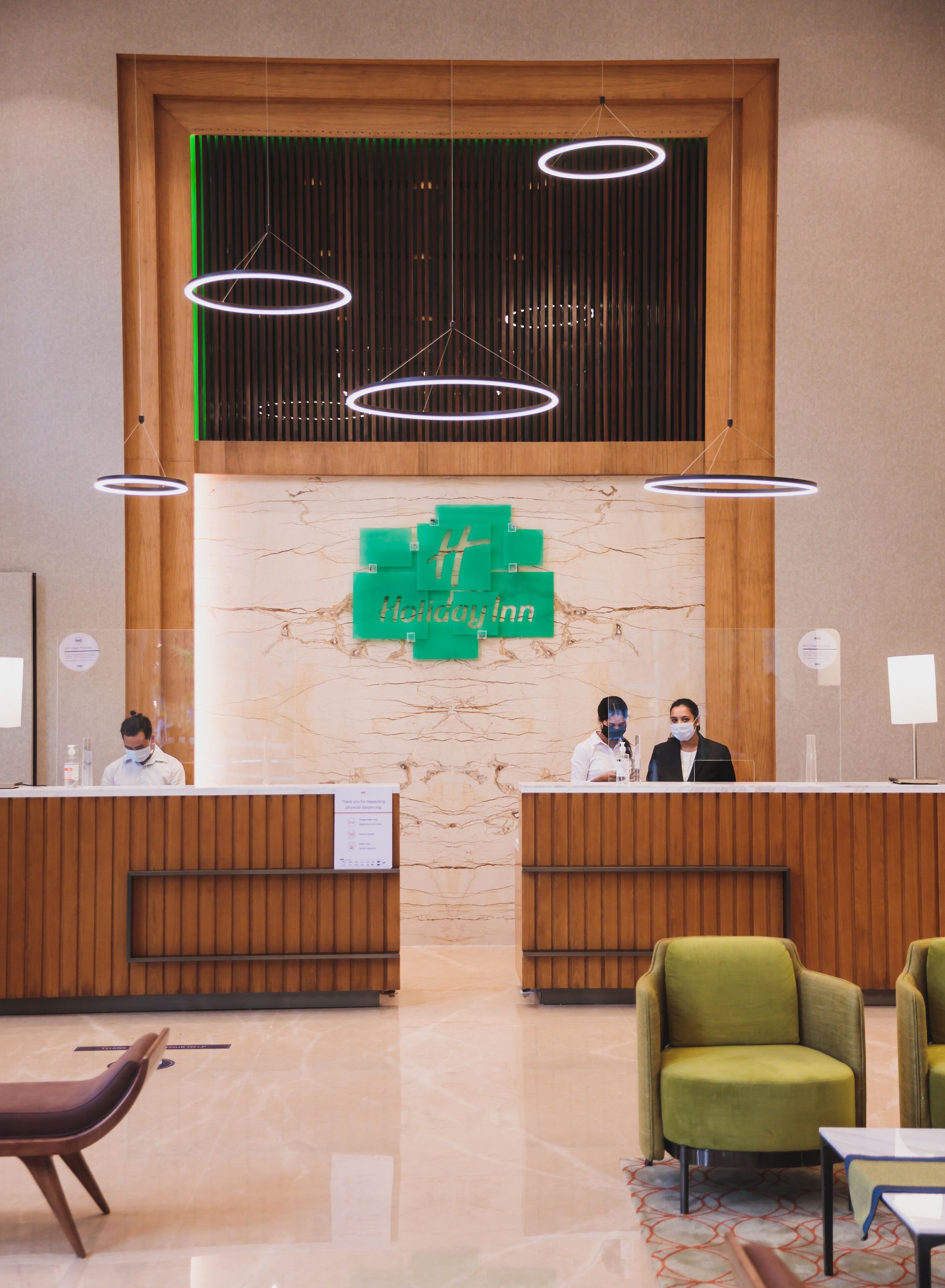 A hotel reception area with a wooden reception desk, a green logo, and modern circular light fixtures.