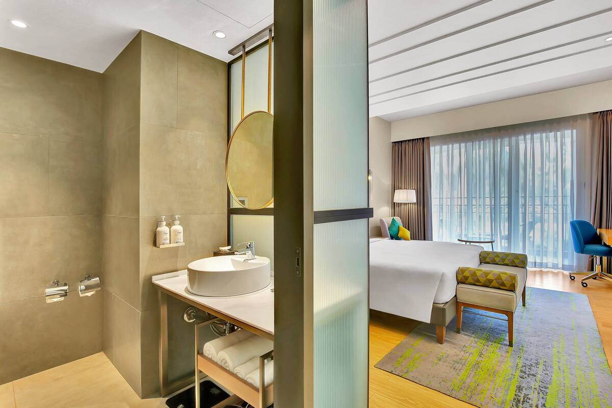 A modern hotel room with a large bed, a round mirror, a built-in sink, and a seating area.