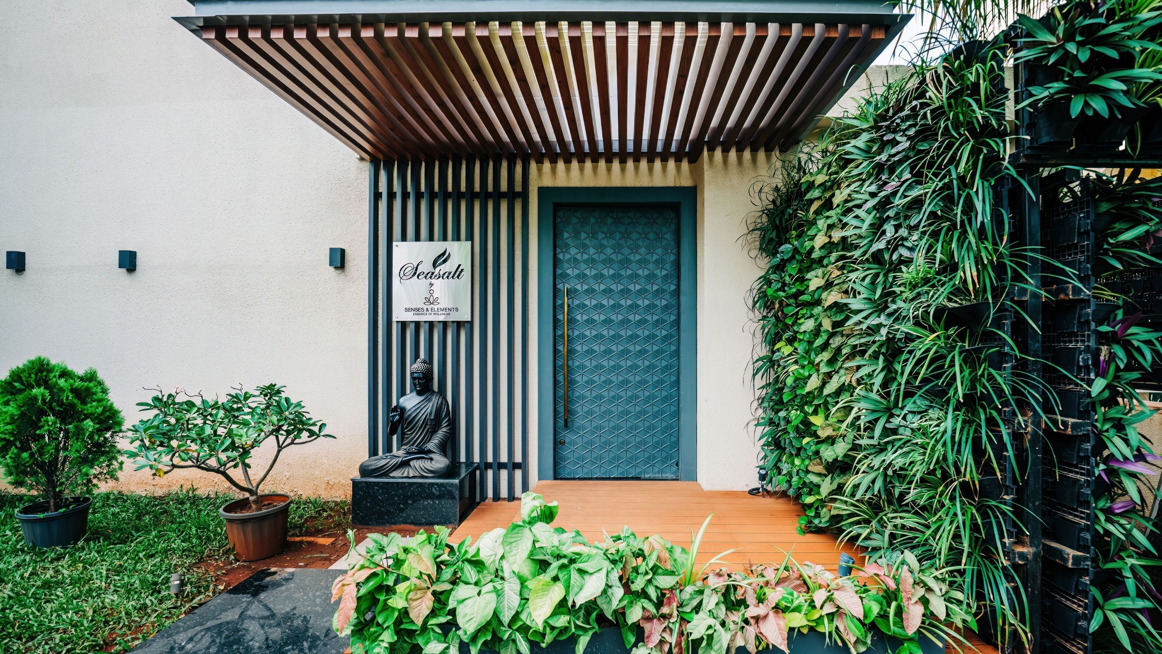Un edificio moderno con una entrada de pérgola de madera, una estatua de Buda y un letrero que dice "Seasalt Senses & Elements Essence of Wellness".