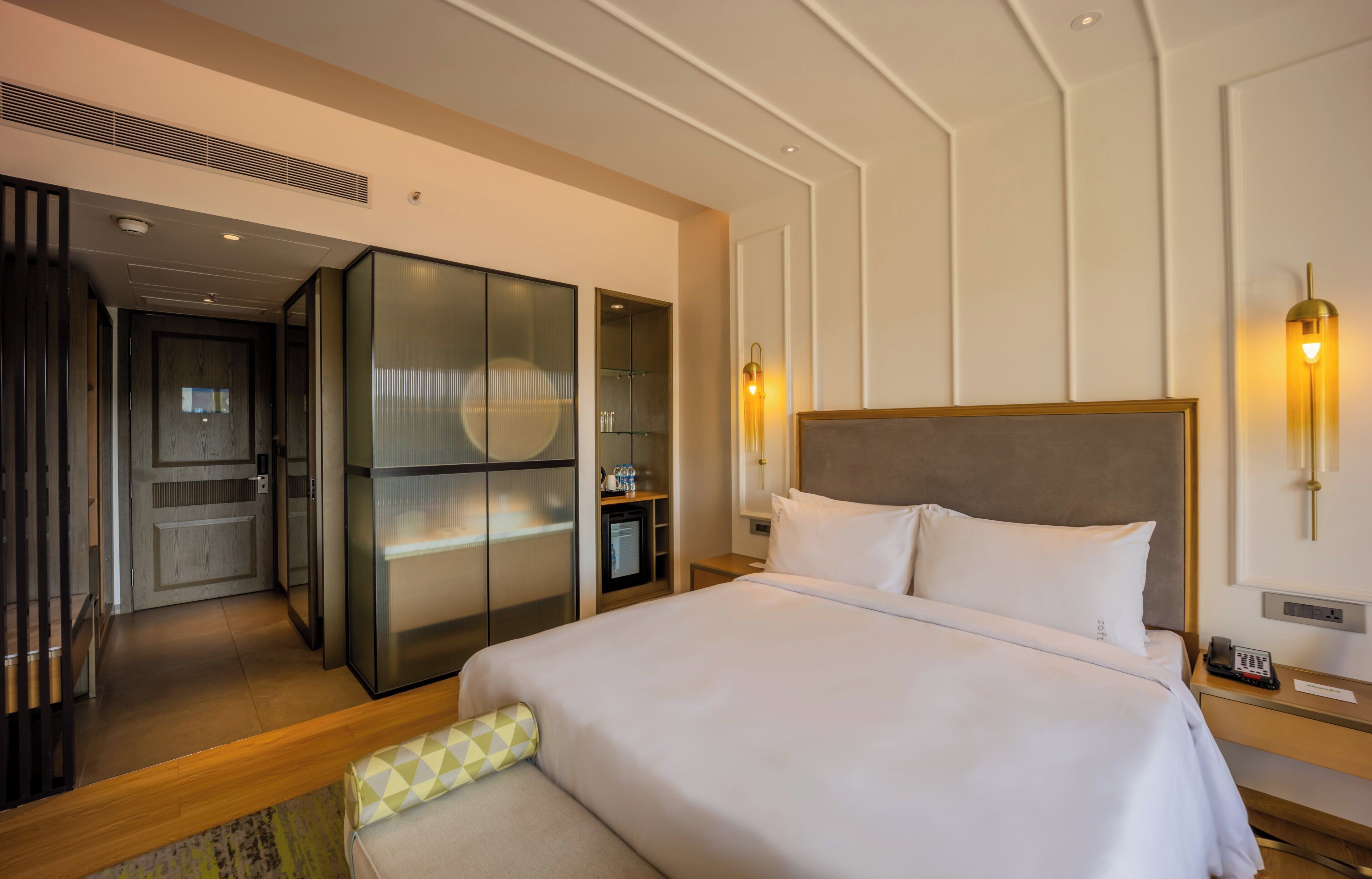 A hotel room with a large bed, a wardrobe, a minibar, and a desk with a phone.
