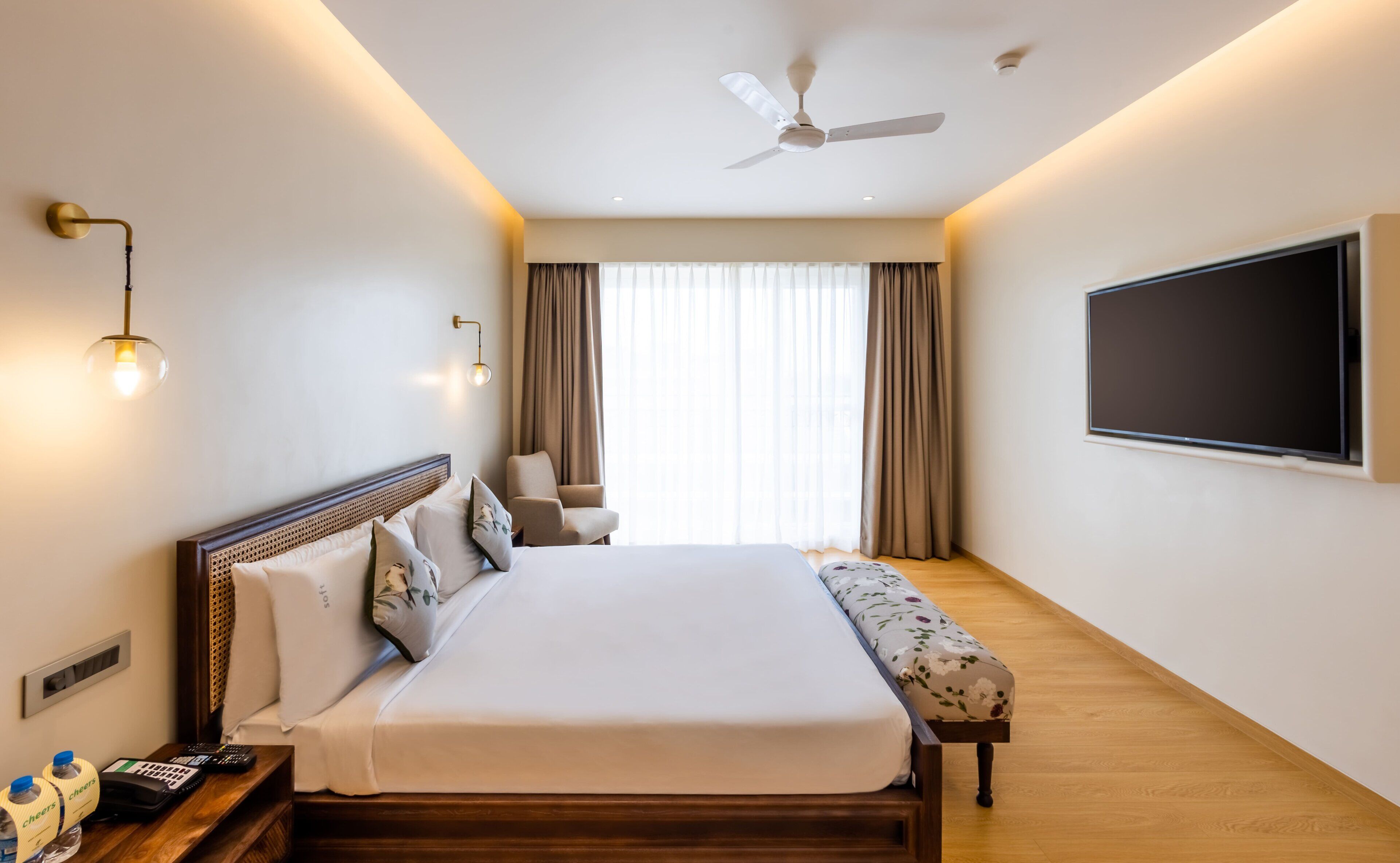 A hotel room with a large bed, bedside table, lamp, ceiling fan, and a TV mounted on the wall.