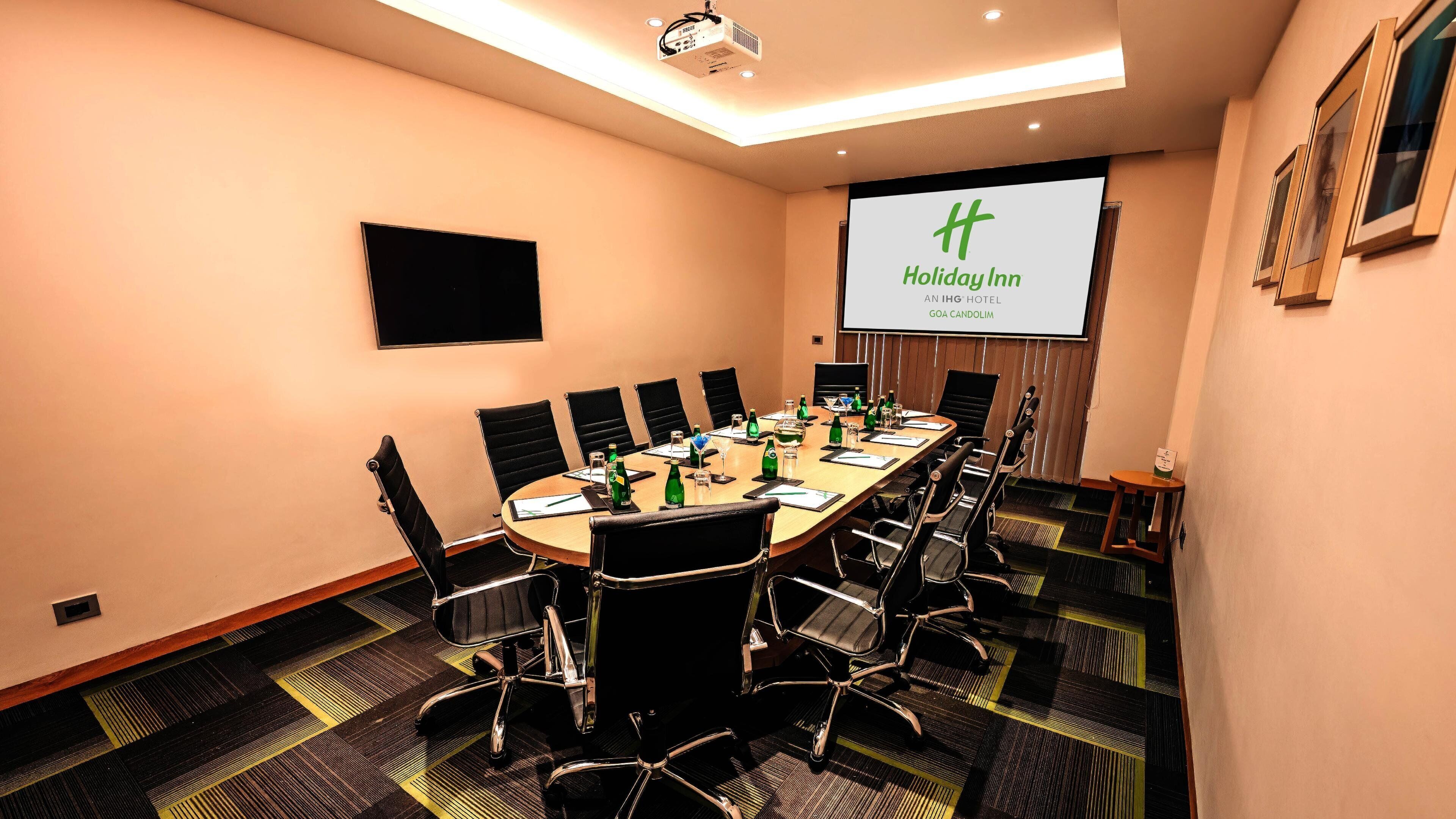 A conference room with a large table, chairs, a projector screen, and a screen displaying the Holiday Inn logo.