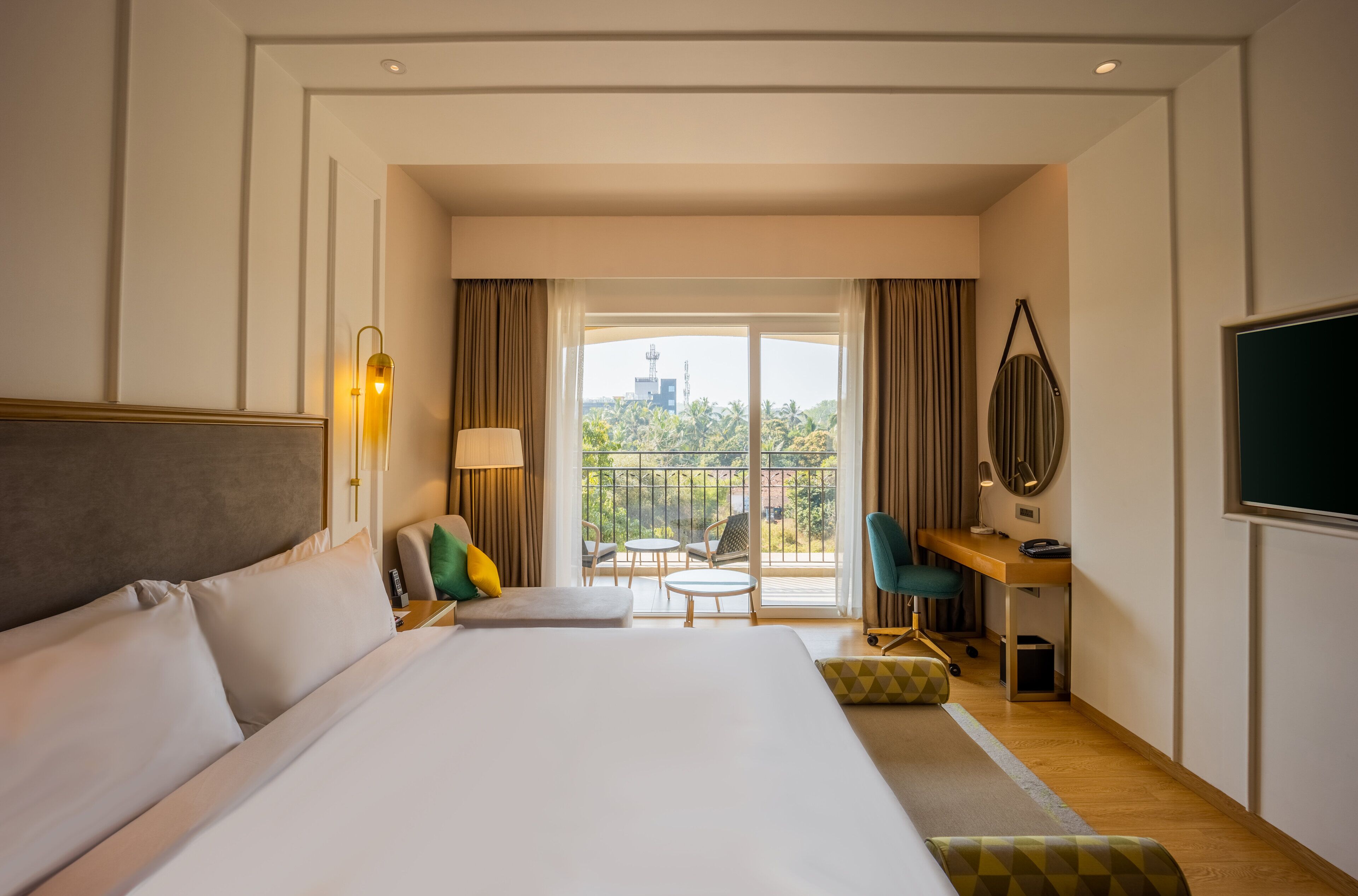 A hotel room with a large bed, a desk with a chair, a TV, and a balcony with a view of greenery and buildings.