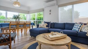 Una sala moderna con un sofá azul, sillas de madera, una mesa de centro redonda y grandes ventanas que ofrecen vistas a la vegetación.