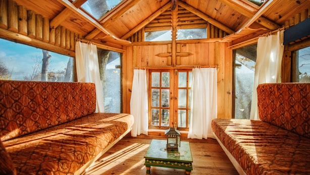 Une pièce chaleureuse aménagée dans un chalet en rondins, avec deux canapés à motifs, une petite table sur laquelle est posée une lanterne, et de grandes fenêtres ornées de rideaux.