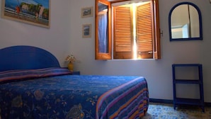 Un dormitorio con cama, ventana con contraventanas de madera, espejo y un cuadro en la pared.