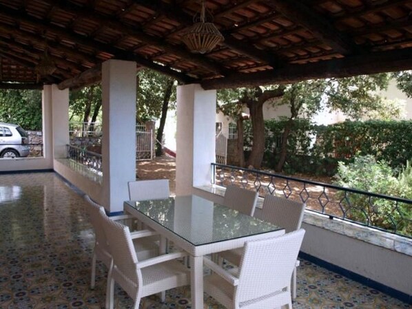 An outdoor patio with a glass-top table and white wicker chairs, surrounded by a tiled floor and a wooden ceiling. - Villa Novella (Acquafredda)