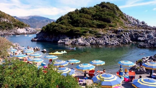 Zona costera con terreno rocoso, cielo despejado y numerosas sombrillas de playa.