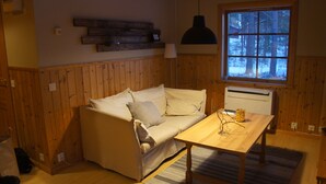 Una habitación acogedora con un revestimiento de madera, un sofá beige con cojines, una mesa de madera y una ventana con vistas a árboles.
