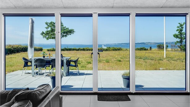 Een moderne kamer met grote schuifdeuren die uitkomen op een terras. Het terras heeft een tafel met stoelen, een potplant en uitzicht op de zee en heuvels in de verte.
