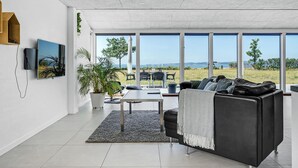 Een ruime woonkamer met een groot raam met uitzicht op zee, een zwarte leren bank, een flatscreen televisie aan de muur en een kamerplant.