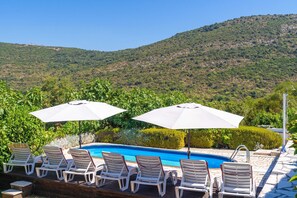A poolside area with lounge chairs and umbrellas, surrounded by lush greenery and a hilly landscape.