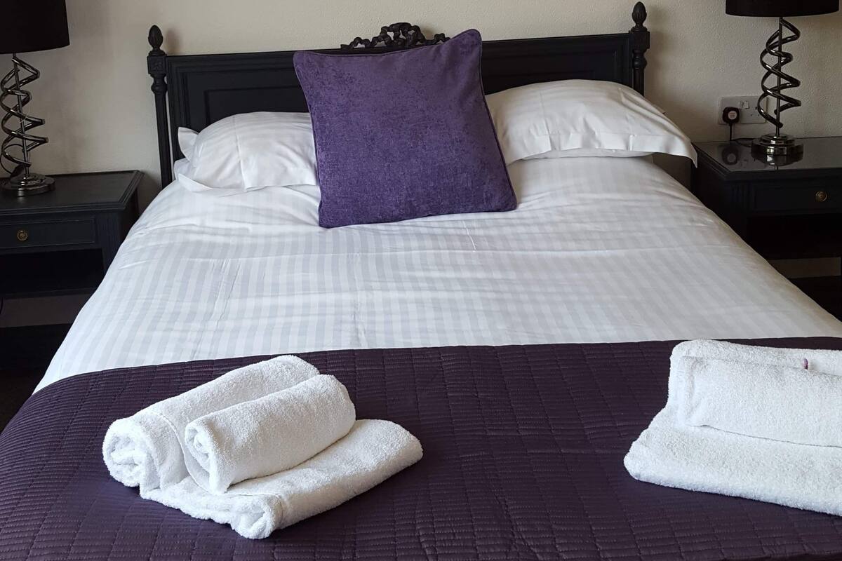 A neatly made bed with white linens, a purple pillow, and two white towels folded on the bed.