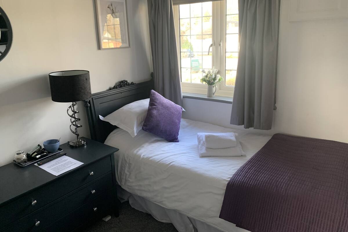 A neatly arranged hotel room with a bed, a bedside table with a lamp, a small desk with a cup and papers, a window with curtains, and a framed picture on the wall.