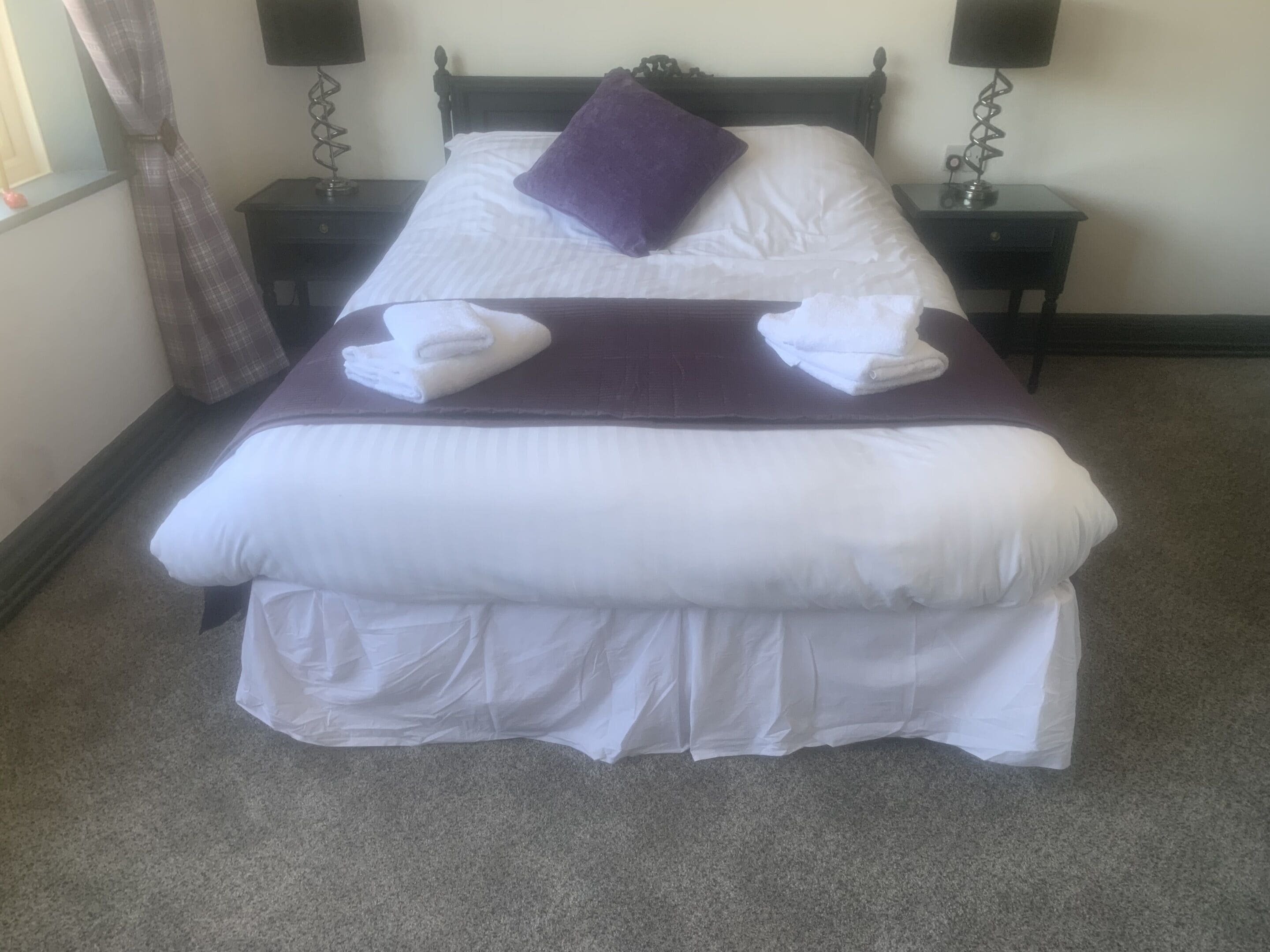 A neatly made bed with white linens, a purple pillow, and two folded white towels.