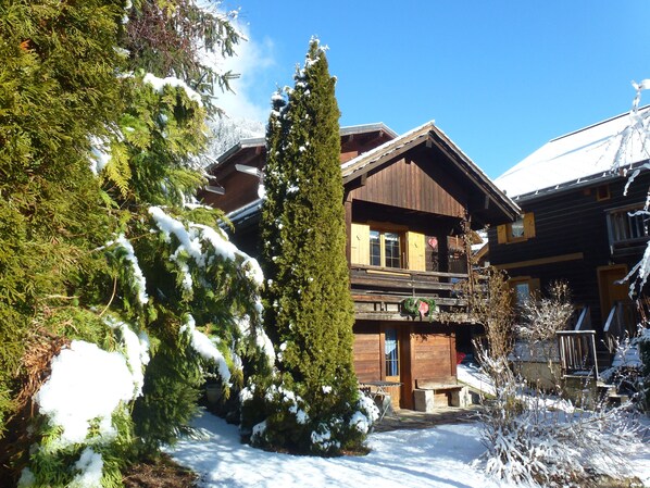 A wooden chalet with a balcony, surrounded by snow-covered trees and a clear blue sky.