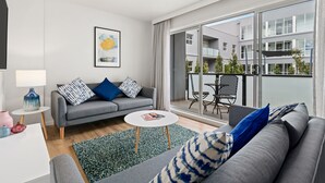 A modern living room with a grey sofa, a small round table, and a balcony with a view of other buildings. - Modern and Spacious Two Bedroom Apartment (Burwood)