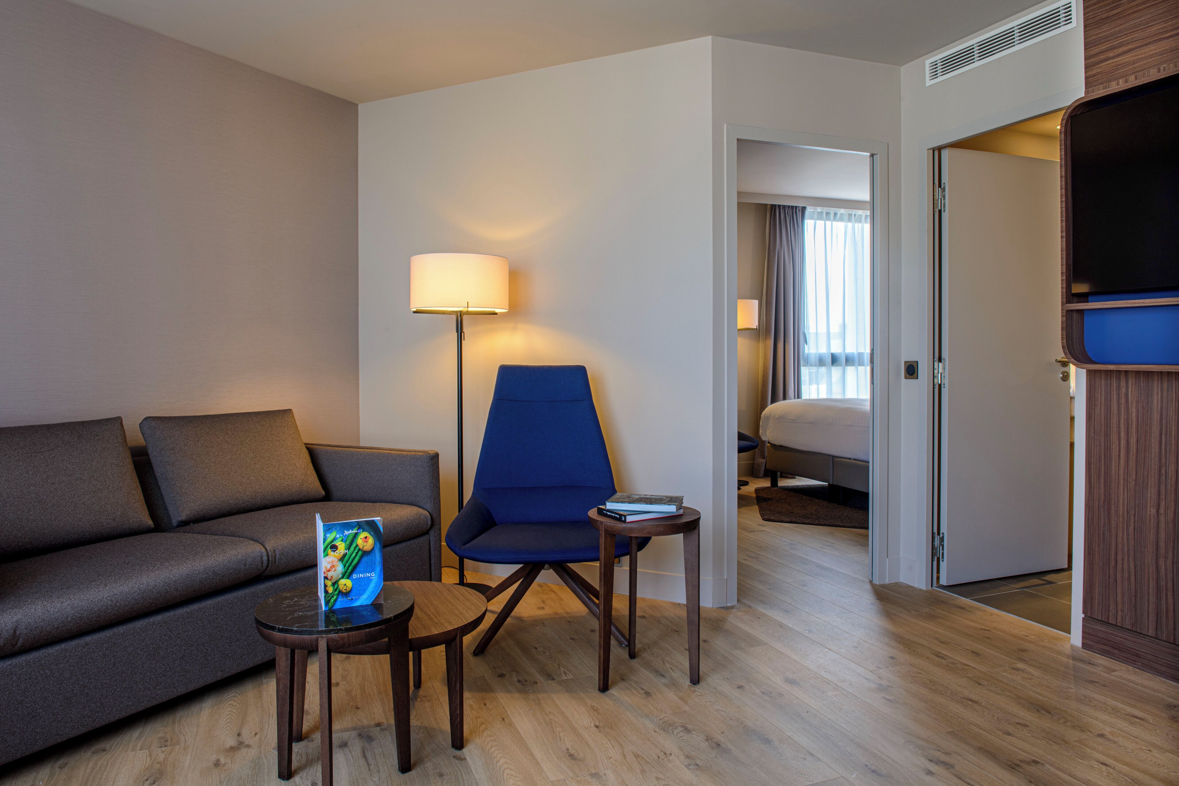A modern hotel room with a sofa, a blue armchair, a small table with a book, a lamp, and a television.