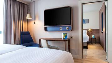A hotel room with a large bed, a desk with a chair, a TV mounted on the wall, and a door leading to another room.