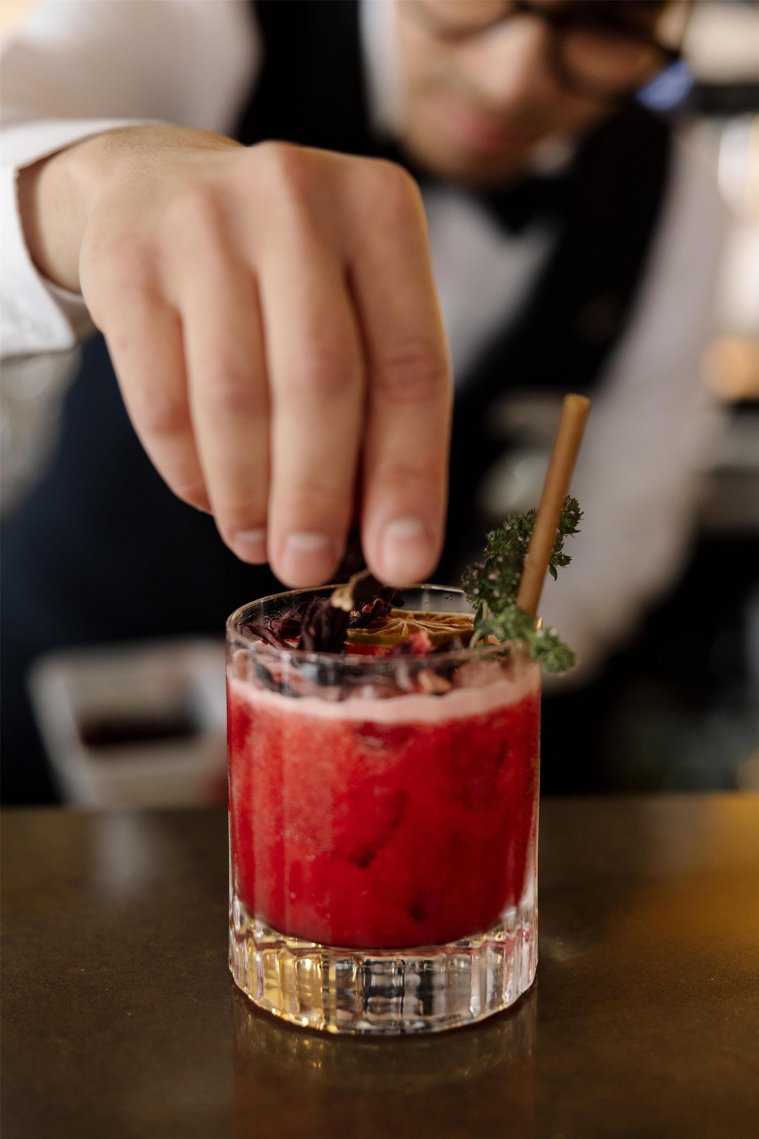 Eine Hand platziert einen Strohhalm in ein Glas mit einem roten Getränk, Eiswürfeln und einer Garnitur.