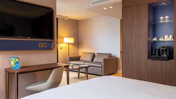 A hotel room with a bed, a desk, a chair, a sofa, a TV, a lamp, and a coffee machine.