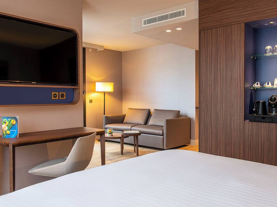 A hotel room with a bed, a desk, a chair, a sofa, a TV, a lamp, and a coffee machine.