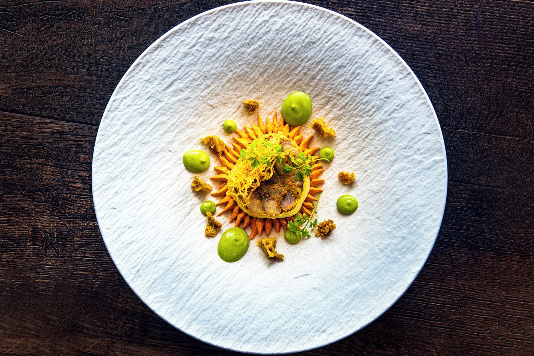 A round white plate with a central dish, surrounded by green spheres and shredded elements.