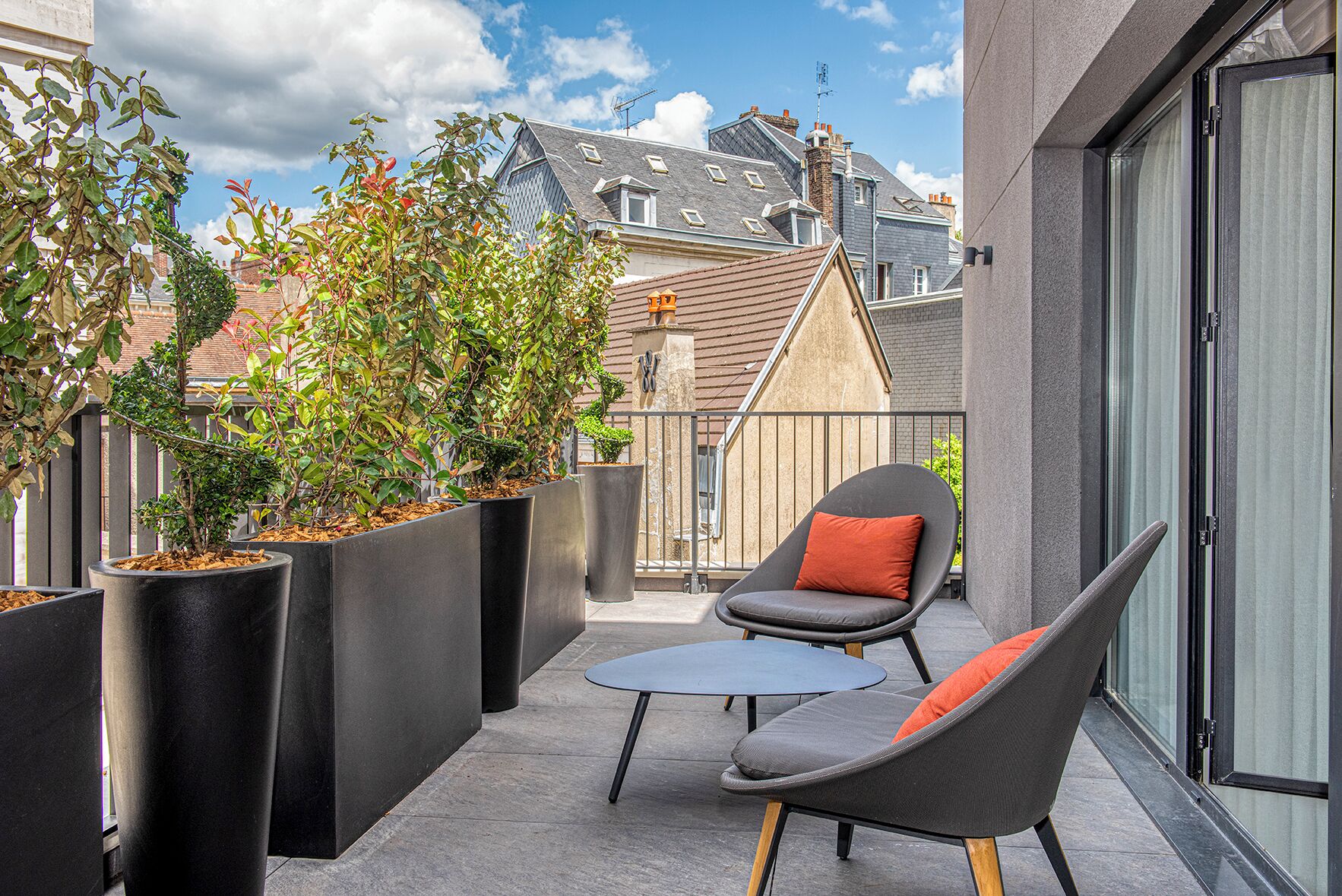 A balcony with modern outdoor furniture, large planters with shrubs, and a view of neighboring buildings.