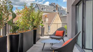 A balcony with modern outdoor furniture, large planters with shrubs, and a view of neighboring buildings.