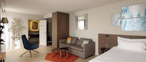 A modern hotel room with a bed, a sofa, a chair, a small table, and a painting on the wall.