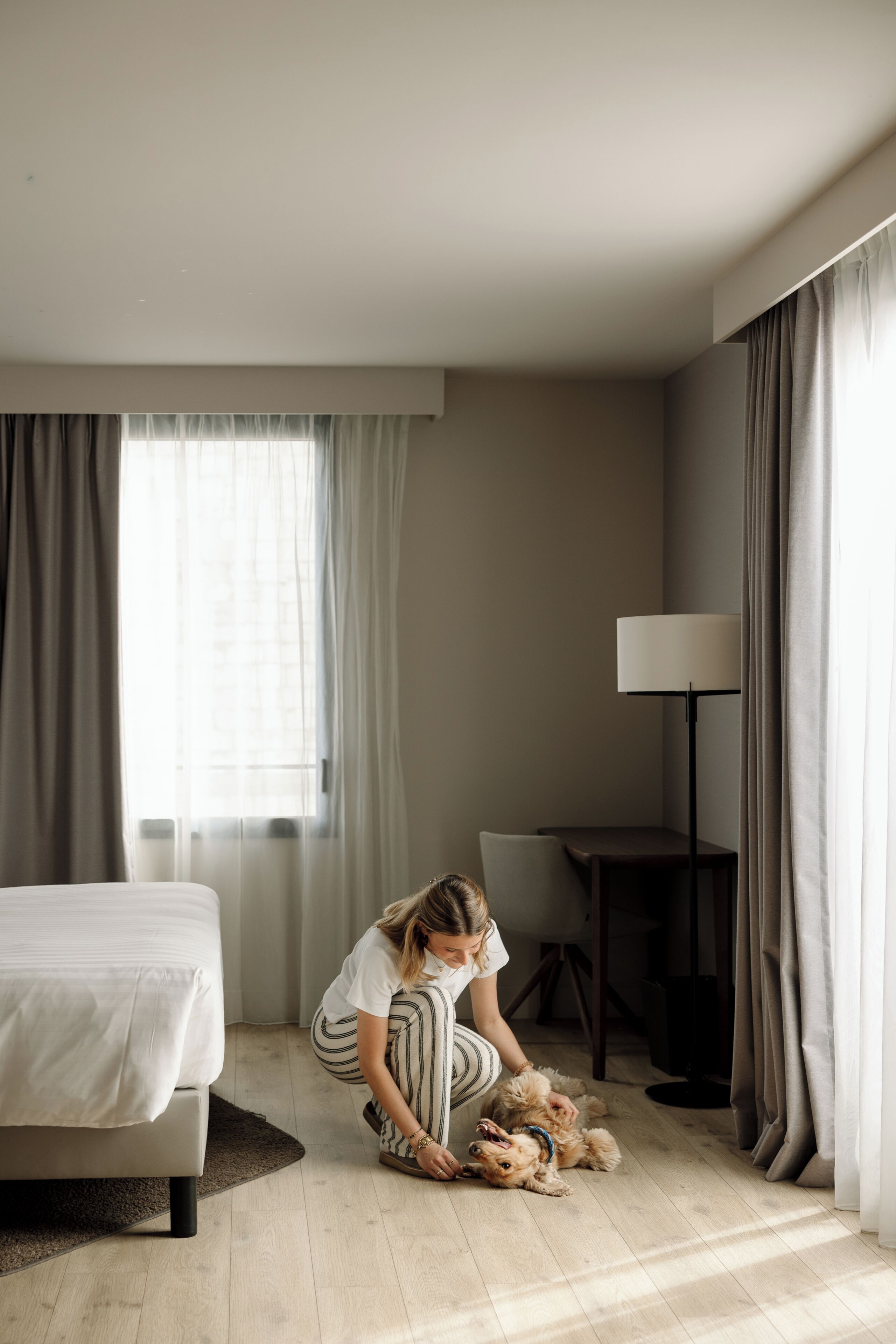 A hotel room with a bed, a desk, a lamp, and a window with curtains.