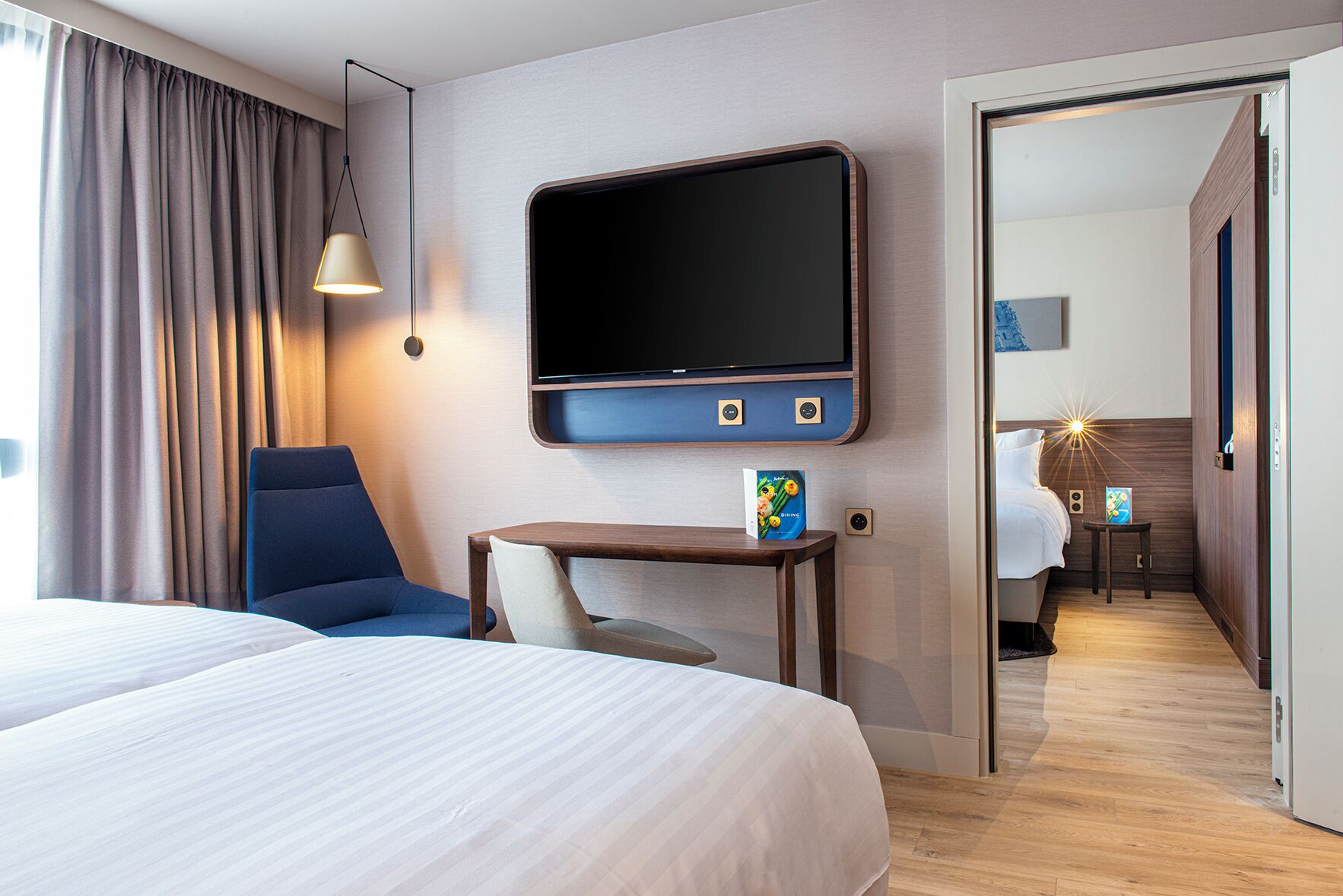 A hotel room with a bed, a desk, a chair, a TV, and a door leading to another room.