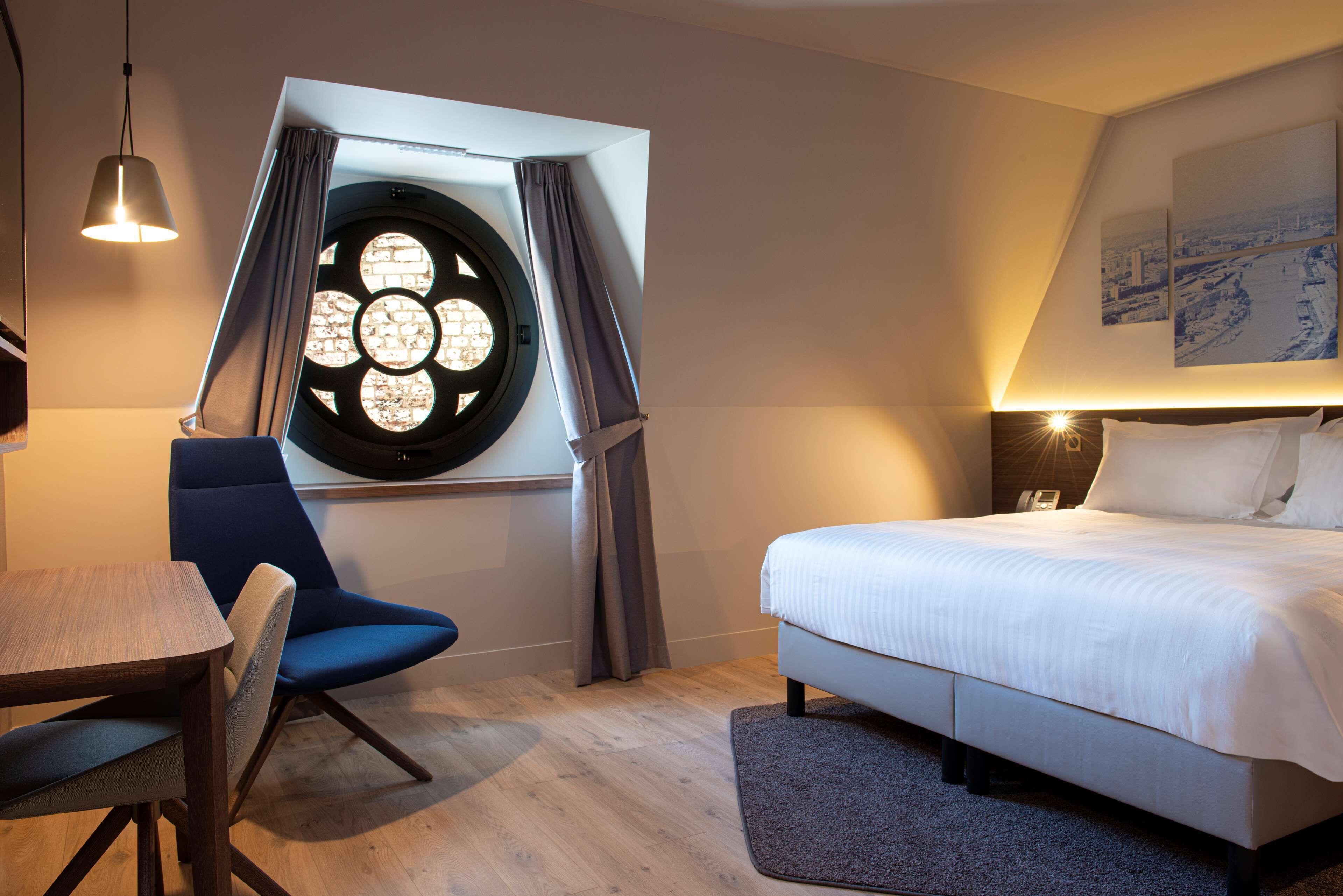 A modern hotel room with a large bed, a dining table, a blue chair, a unique circular window, and a cityscape painting on the wall.