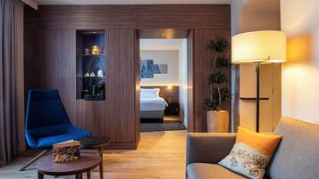 A modern hotel room with a blue armchair, a grey sofa, a small table with a book, a bedside lamp, and a bed with a nightstand.