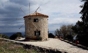 A stone windmill with a tiled roof, surrounded by a cobblestone path and flowering plants. - Potamitis Windmills & Apartments (Zakynthos)