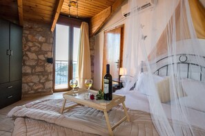 A bedroom with a stone wall, wooden ceiling, a bed with white bedding, a bedside table with a bottle of wine and glasses, and a window with curtains. - Potamitis Windmills & Apartments (Zakynthos)