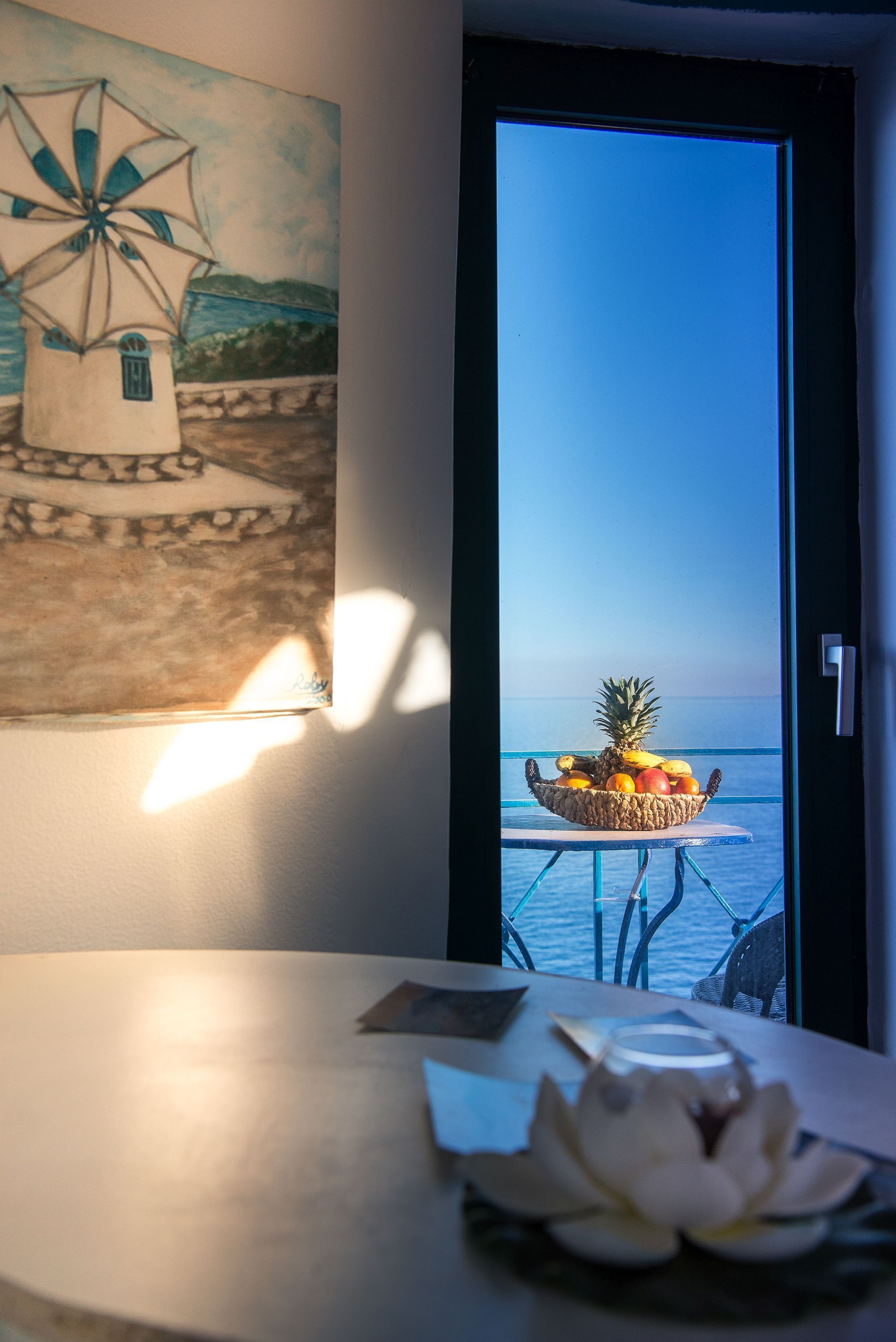 A table with a fruit bowl and a white flower centerpiece, a painting of a windmill, and a view of the sea through an open door.