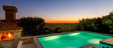 A swimming pool with a stone fireplace and seating area, surrounded by greenery and a view of the sunset.