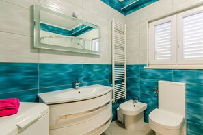 Et badeværelse med et hvidt håndvask, et lille væghængt håndvask, et toilet og en bidet. Et spejl er placeret over håndvasken, og der er et vindue med persienner.