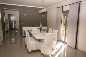 A modern dining area with a white table and chairs, a chandelier, and a view of the outdoors through a sliding door. - Villa Elegio by CorfuEscapes (Corfu)