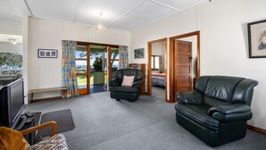 A spacious living area with a large leather sofa, a smaller armchair, and a bench. There is a door leading to a bedroom with a bed and a window. A framed picture hangs on the wall, and a small table is visible in the corner. - Marina Views - Motuoapa Bach (Motuoapa)
