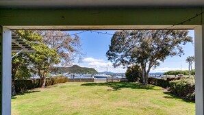 A view from a covered porch with a grassy lawn, trees, and a marina with boats in the background. - Marina Views - Motuoapa Bach (Motuoapa)
