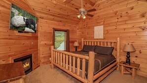 Un dormitorio de cabaña de madera con una cama, chimenea, ventilador de techo y un televisor mostrando un oso.