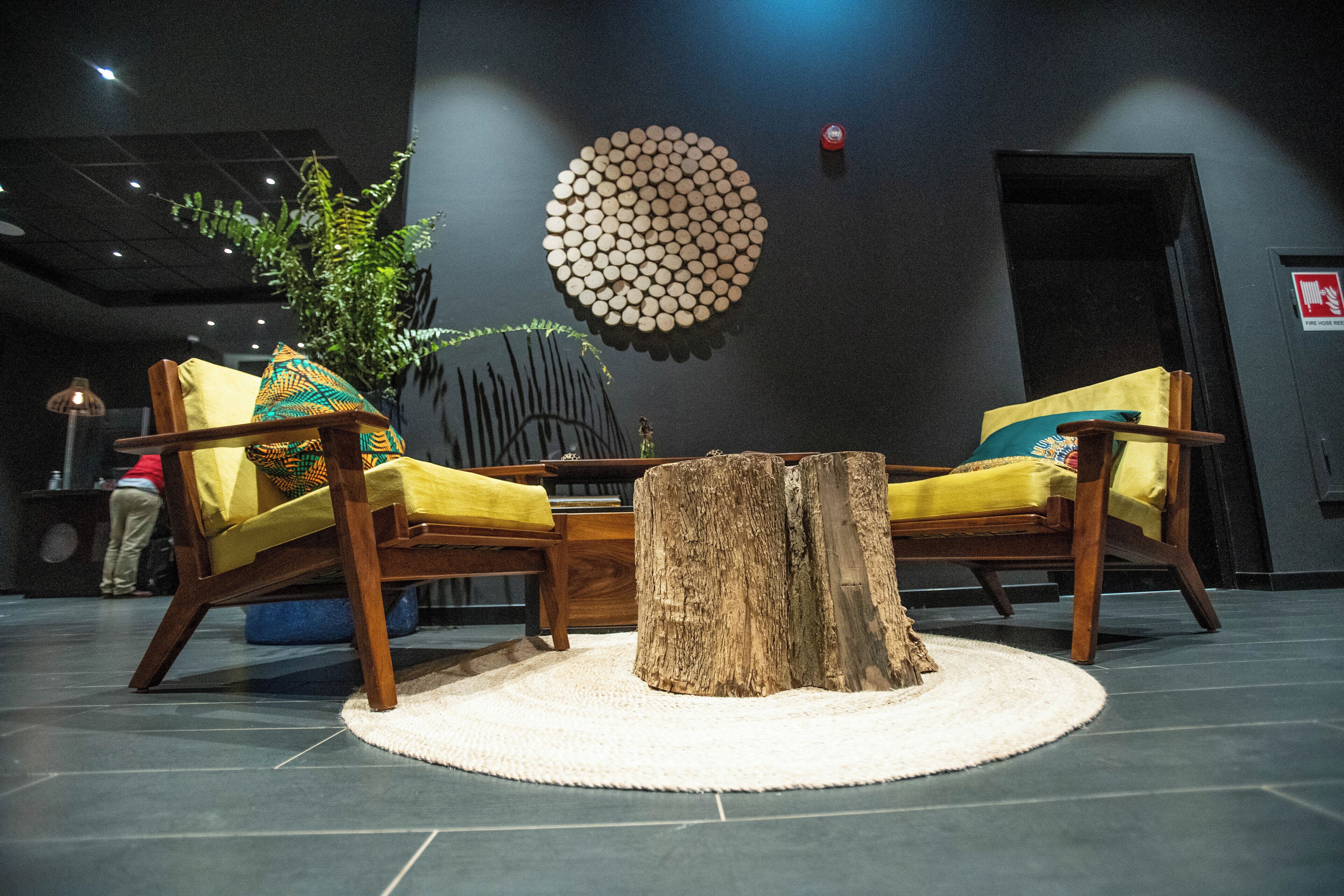 A modern lobby with a round wooden table, two wooden chairs with yellow cushions, a large circular wall decoration, and a potted plant.