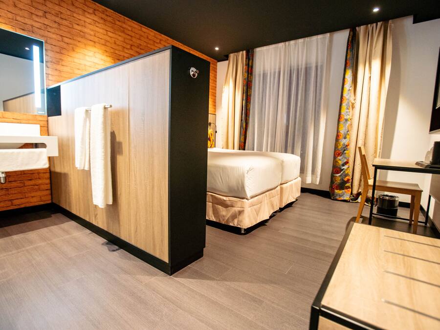 A hotel room with a wooden wall, a bed, a desk, and a bathroom with a sink and mirror.