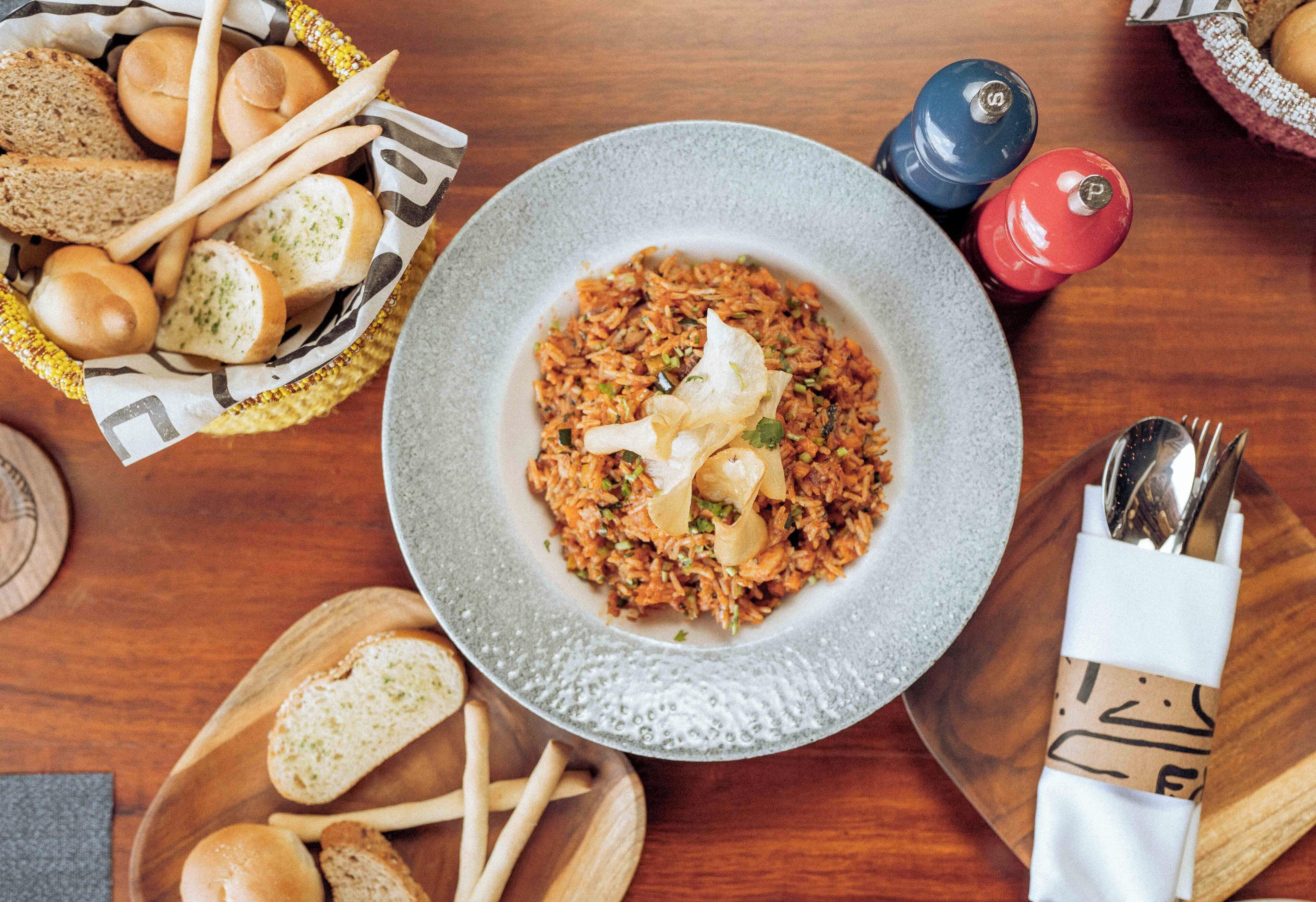 A plate of rice with vegetables and a dollop of sauce, surrounded by bread and condiments on a wooden table.