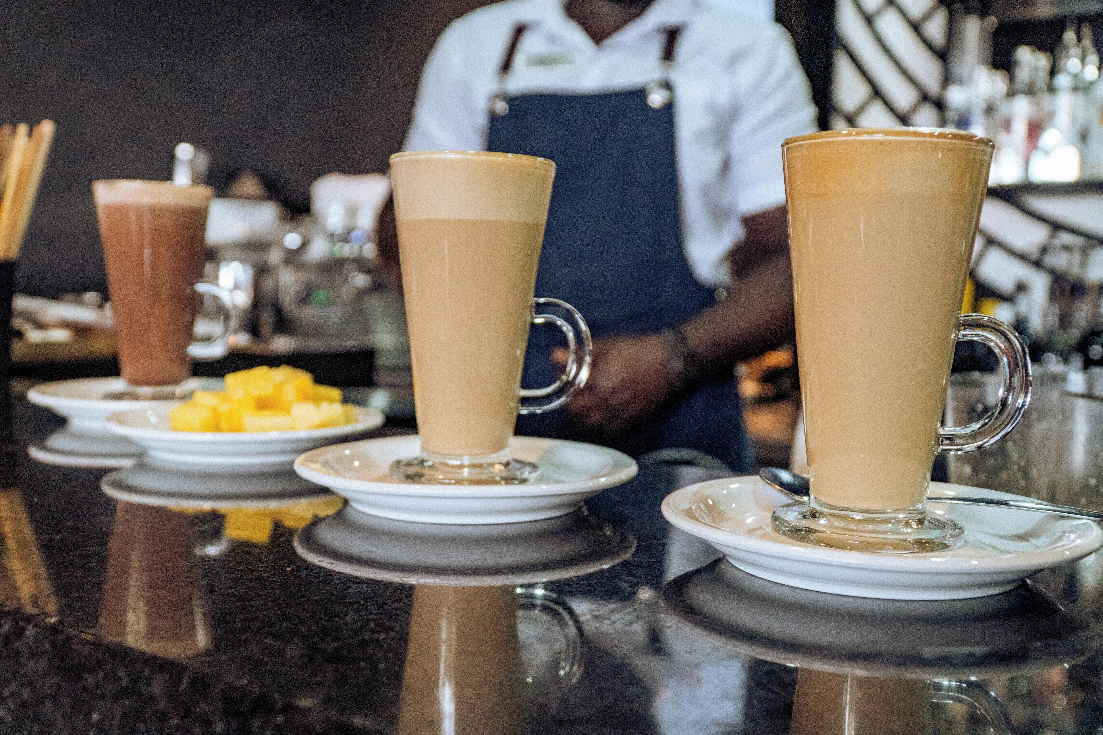 A bar setting with tall glasses of beverages on white saucers, a person in an apron, and a counter with various items.