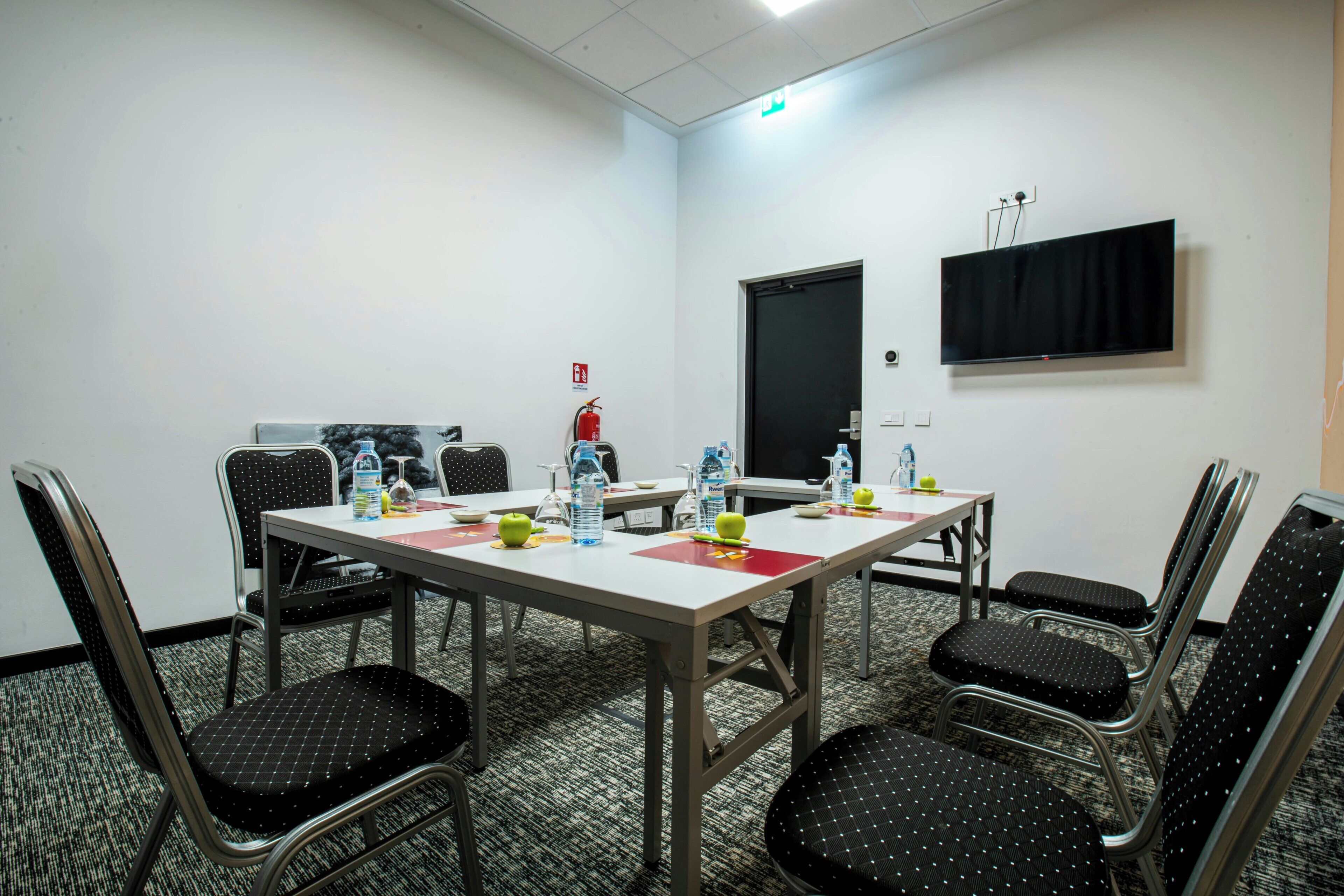 Une salle de conférence équipée d’une table, de chaises, d’un extincteur, d’une porte et d’un téléviseur fixé au mur.