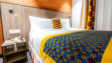 A hotel room with a wooden headboard, a bed with white bedding and a colorful patterned blanket, a bedside table with a phone and a lamp, and a curtain with a red and yellow pattern.