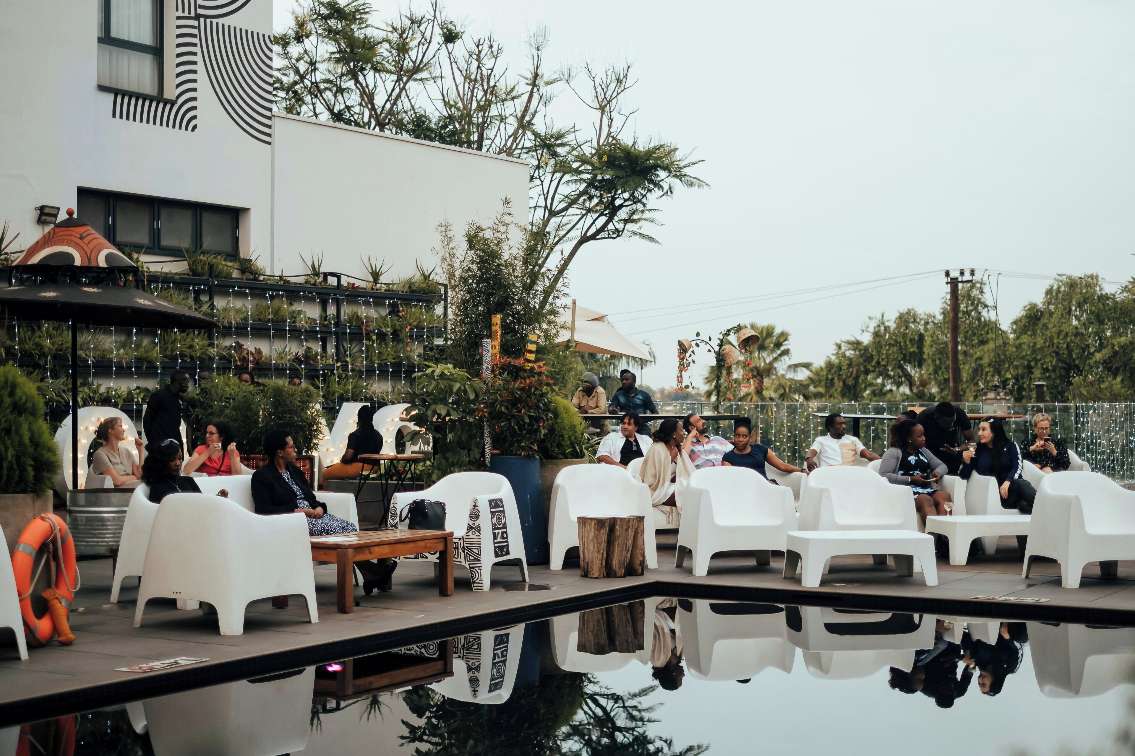 An outdoor seating area with white plastic chairs and tables, surrounded by greenery and a pool.